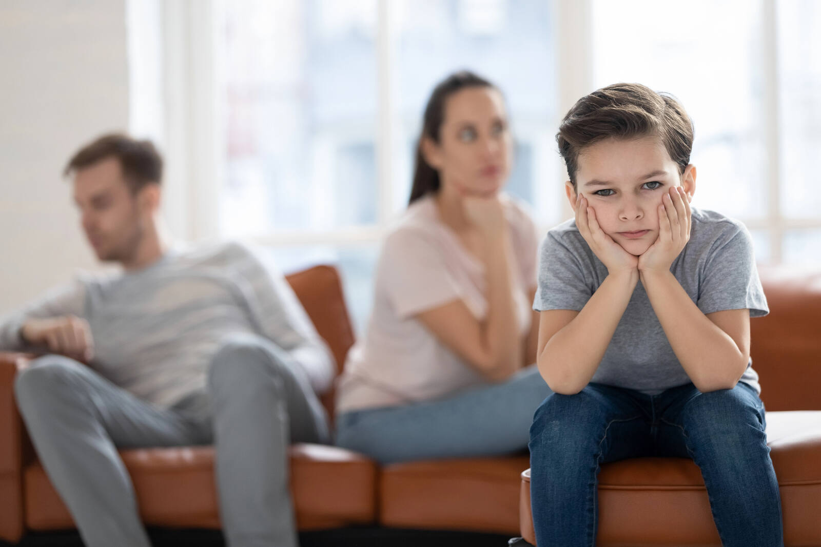 An unhappy young boy sits in the foreground while his parents face away from each other on a couch behind him.