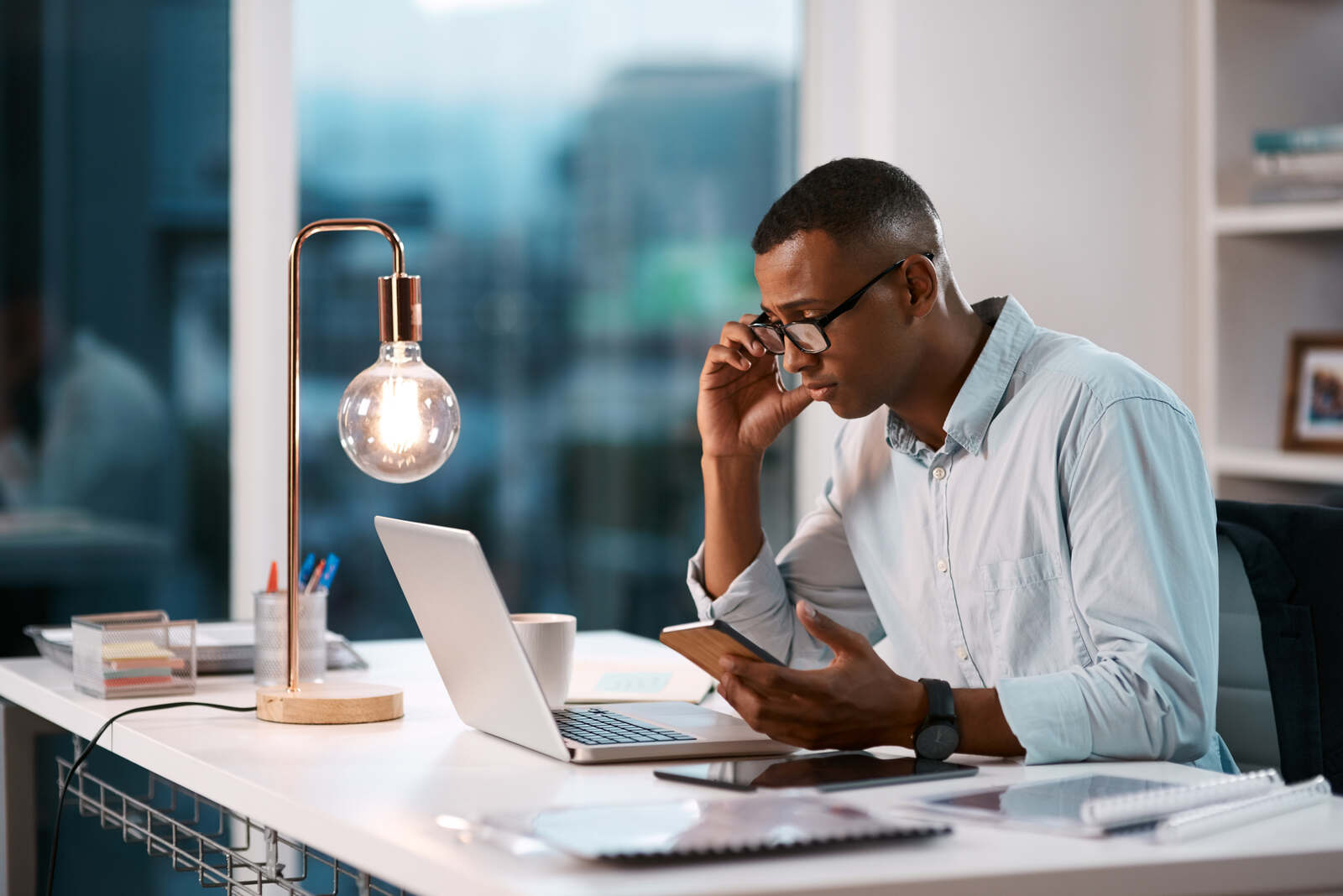A young professional contemplating ideas as he uses his laptop and mobile phone in an office during the evening.