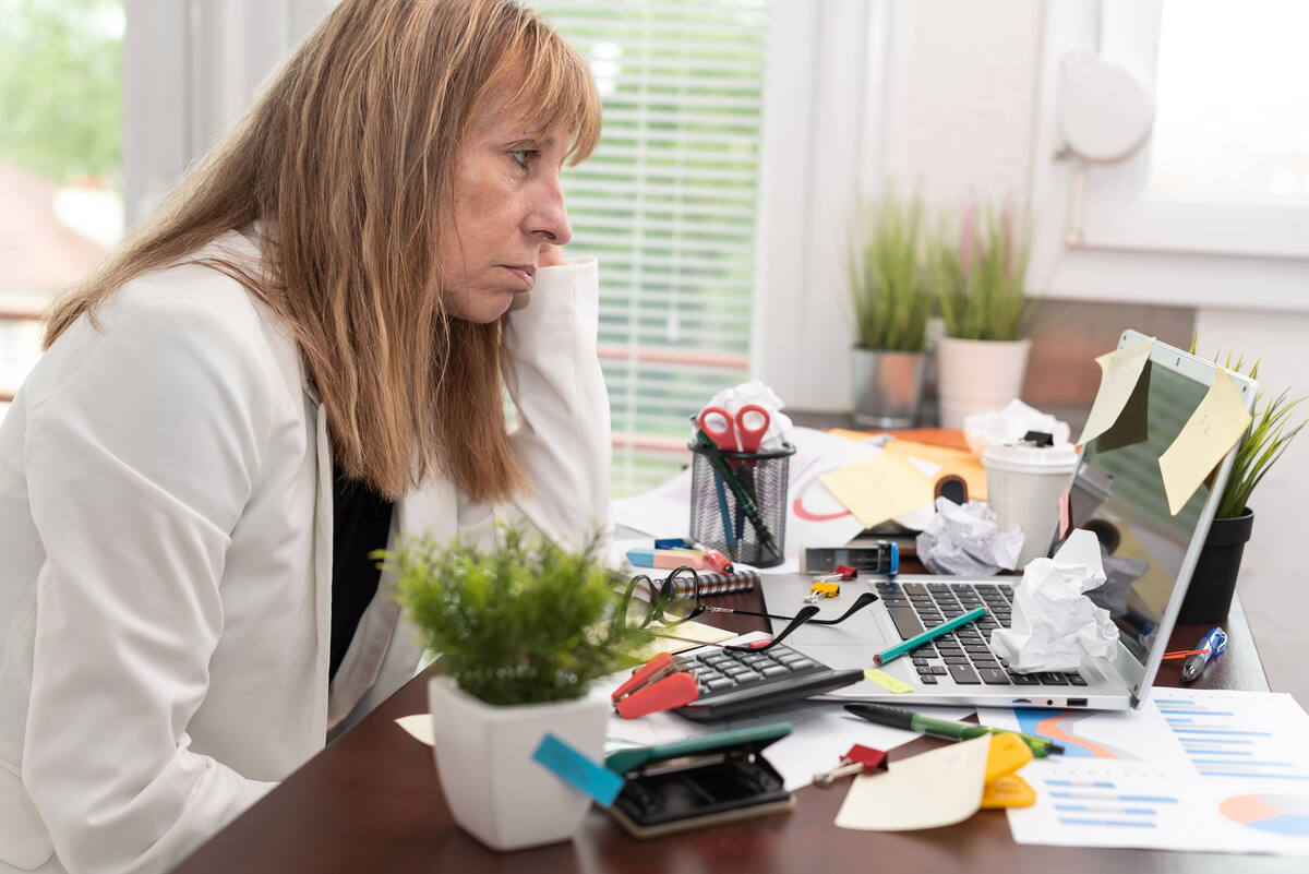 An unhappy looking professional woman stares at her laptop screen; her desk covered in crumpled papers and sticky notes.