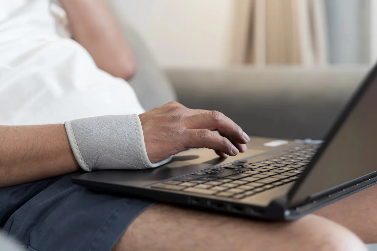 Man wearing a wrist brace after an injury, typing on a laptop while sitting on his couch