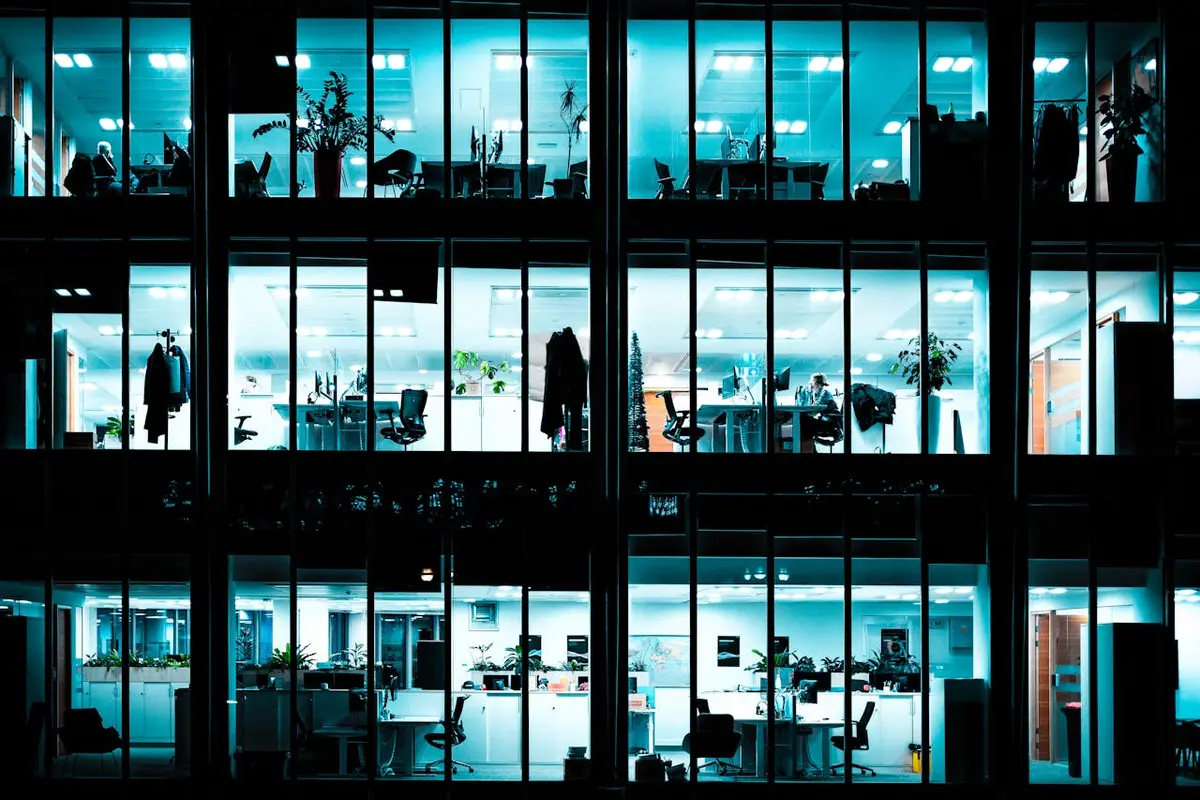 Looking into the windows of a brightly lit office building at night, showing worker desks and offices.