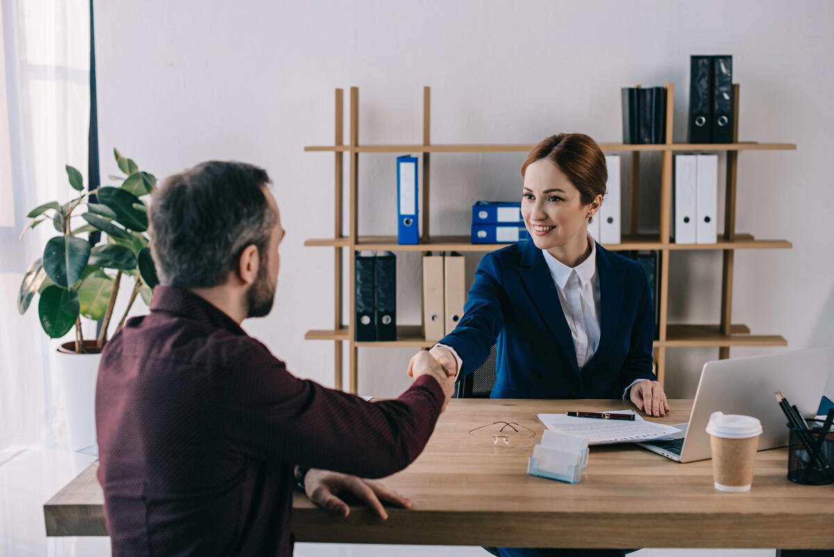 A female lawyer in a blue suit engaging in a handshake with a man in a burgundy shirt. Set in a bright office with a wooden table, potted plant, and organized shelves, it conveys a positive, collaborative mood ideal for business, meetings, or successful negotiations.