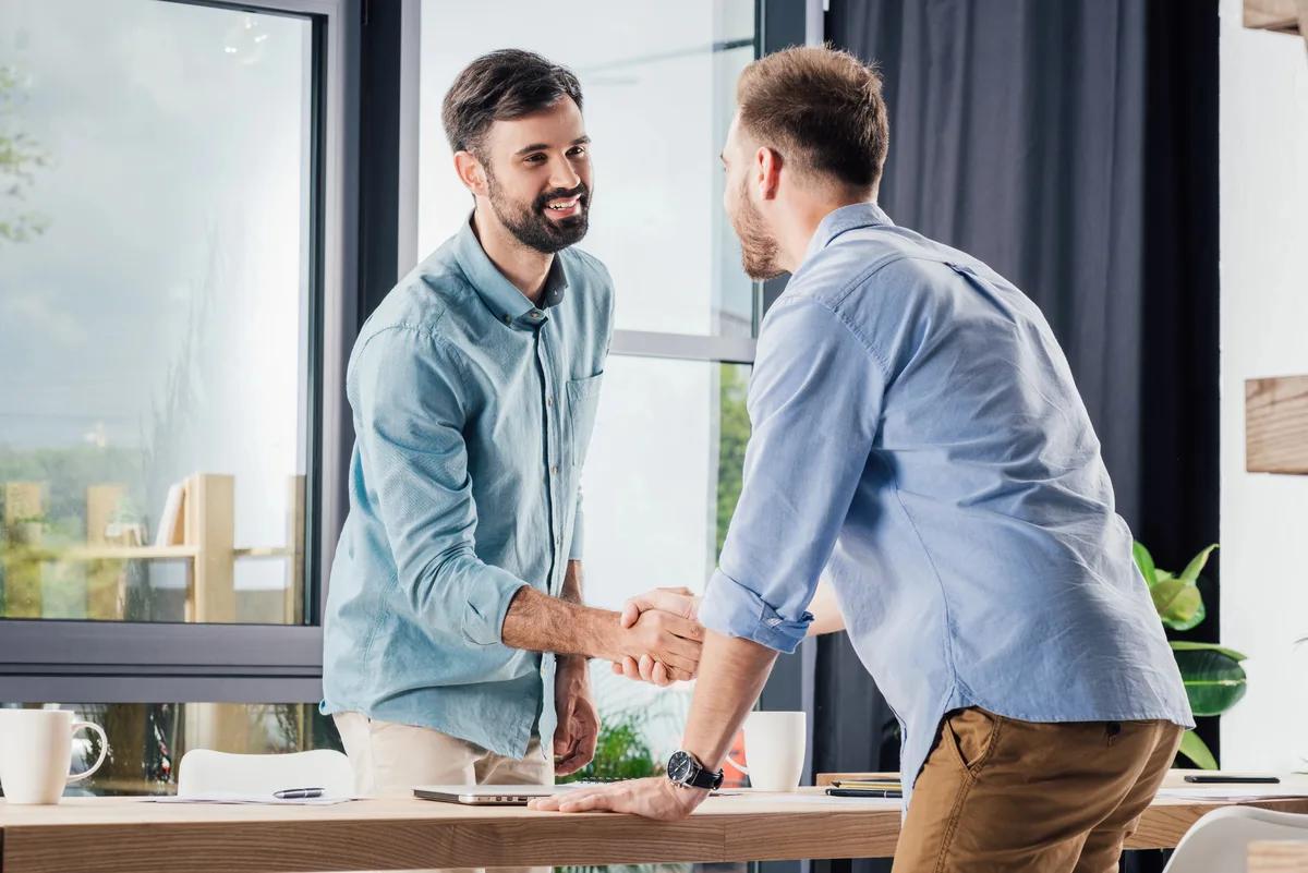 A smiling lawyer shakes hands with a client across a meeting table in a bright office, with coffee mugs, a laptop and papers on the table.