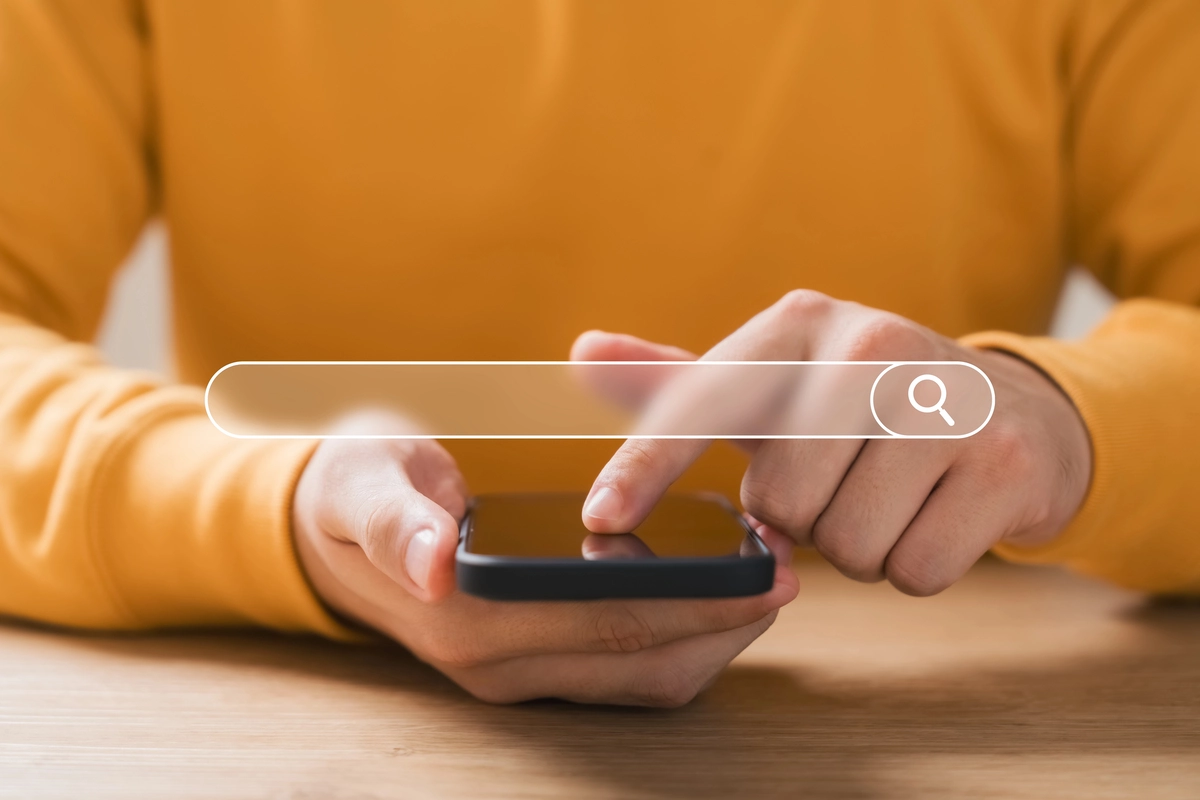 A user in a bright yellow sweater taps on the search bar on their mobile phone.