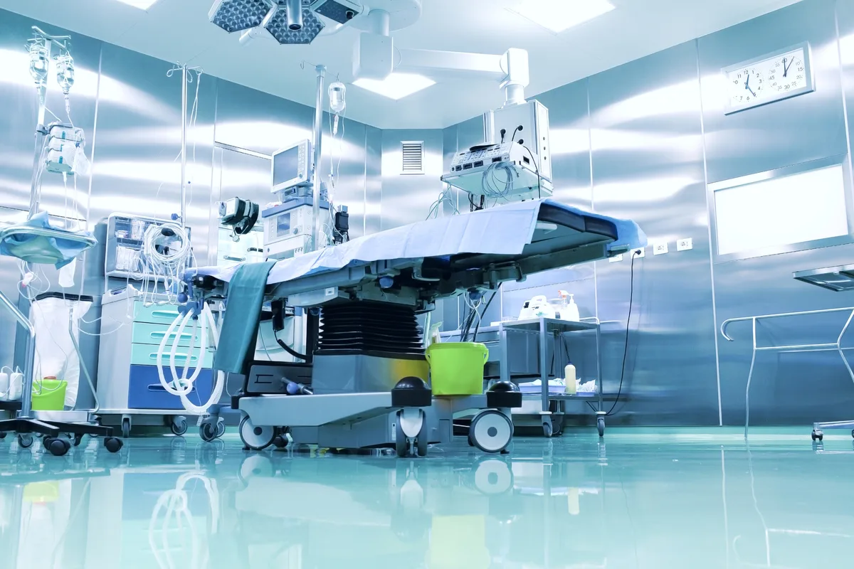 Empty hospital operating room with a surgical table under overhead surgical lights, surrounded by anesthesia equipment, monitors, and IV stands