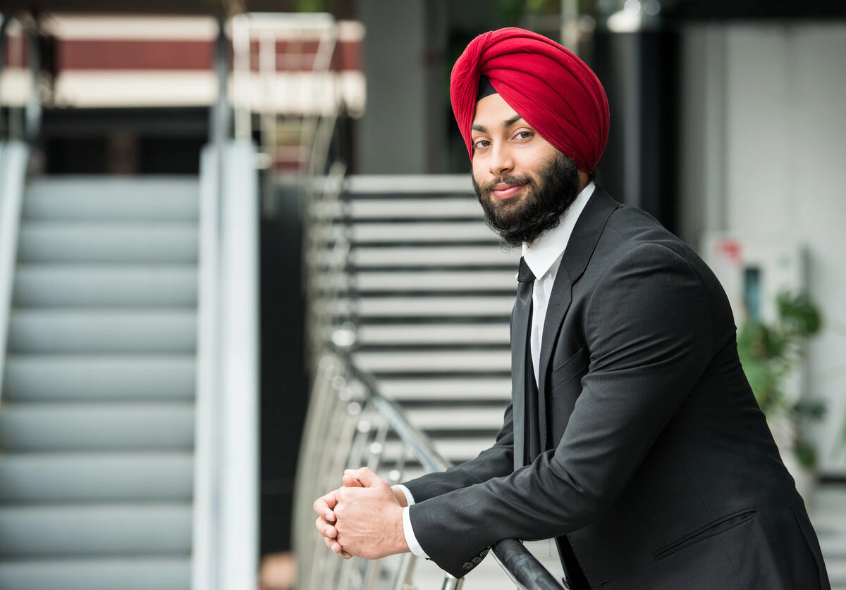 A well-dressed man in a bright red turban leans against a railing inside an office building.