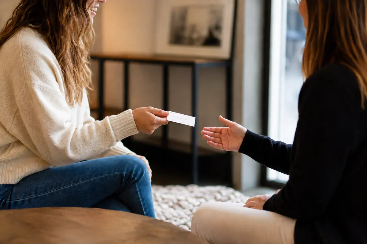 Two young women sit in a warm, casual Canadian office style setting as one hands a business card to the other, suggesting a friend recommending a lawyer.