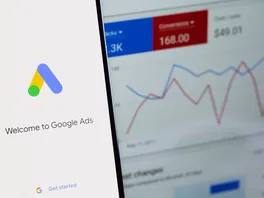 Smartphone showing the Welcome to Google Ads screen in front of a monitor displaying a Google Ads dashboard with clicks, conversions, and a performance chart