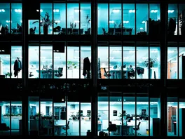 Looking into the windows of a brightly lit office building at night, showing worker desks and offices.